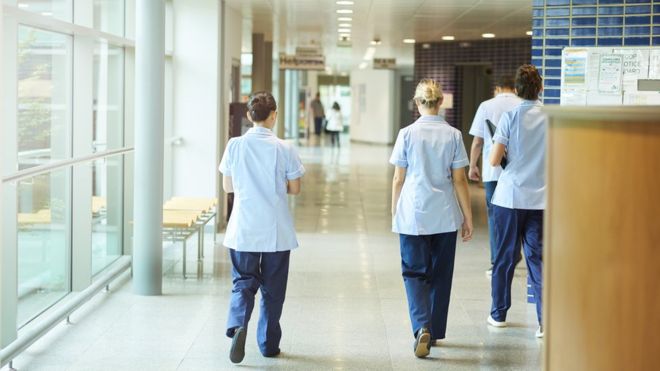 A group of four young trainee nurses including male and female nurses, walk away from camera down a hospital corridor. They are wearing UK nurse uniforms of trousers and tunics.