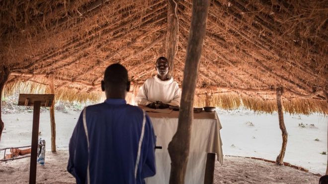 A man and a priest in Southern Sudan stood inside a hut at the refugee camp of Obo The man has his back turned facing the priest as he conducts mass.