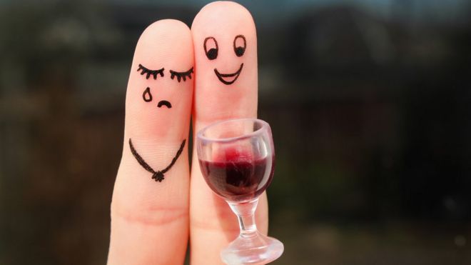 Dedos com desenhos de carinha de feliz e triste perto de uma taça de vinho