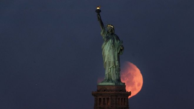 The statue of liberty is seen with pink glowing moon, large around its base