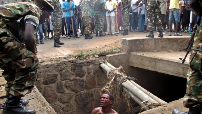 A bloodied man in a pit begs as he is surrounded by soldiers