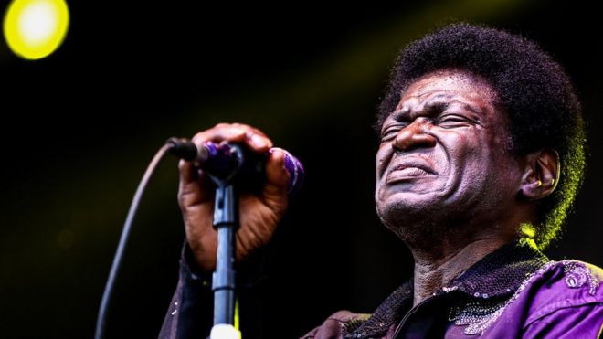 Charles Bradley seen in close-up, eyes closed in front of a stage microphone, wearing a sequined purple coat