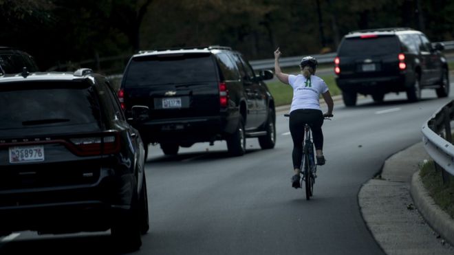 A line of motorcade cars and Juli Briskman on a cycle making a middle finger gesture