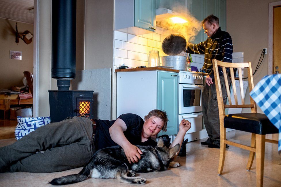 A young man strokes his dog as his father cooks behind him.