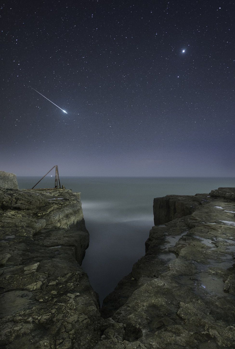 A shooting star flashes across the sky over the craggy landscape