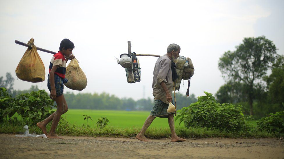 Two generations of a Rohingya refugee family walk with their belongings attached to long sticks across their shoulders