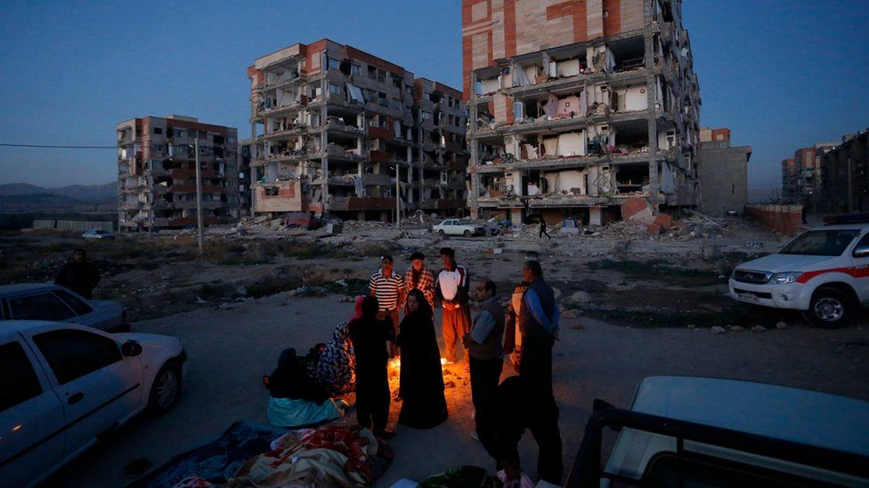 Residentes en Sarpol-e Zahab sentados en torno a una hoguera frente a edificios destruidos.