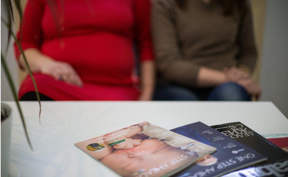 Tetiana y Jana, dos sustitutos, se sientan en una sala de espera frente a una mesa de café con revistas, una de las cuales tiene la cara de un bebé en la portada