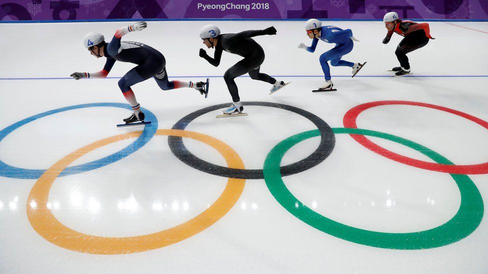 Four male skaters race each other with the Olympic rings image seen in the ice beneath them