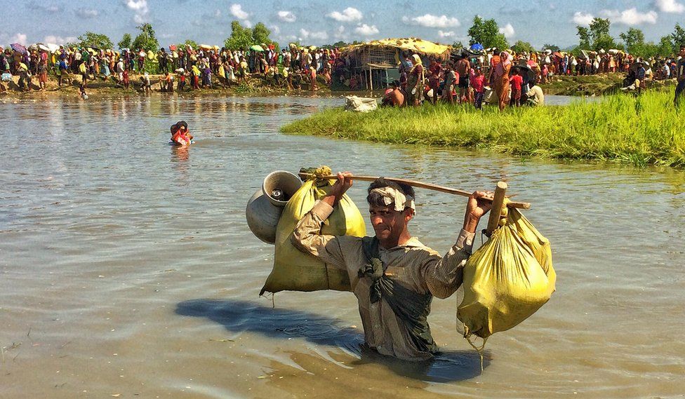 A Rohingya man wading in waist-deep water carries sacks and pots over his head, looking directly at the camera